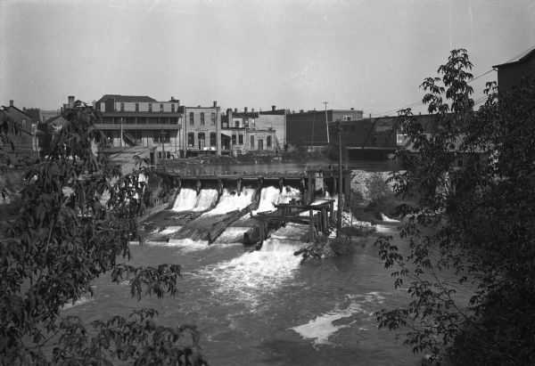 Elevated view of a river, and Fergus Falls buildings in the background. Buildings lining the river have signs that read: "E.T. Bernard," "Hardware," and "Saloon."