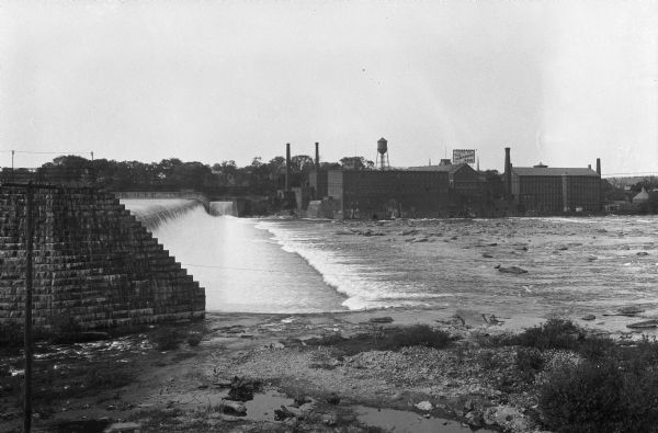 View of a dam, with industrial buildings on the opposite shoreline. A factory building has a sign that reads: "Home of Old Hampshire Bond," and "Hampshire Paper Company."