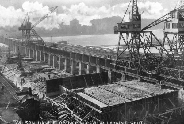 Elevated view of Wilson Dam under construction, looking south. Three large cranes can be seen on the dam over the Tennessee River. Caption reads: "Wilson Dam, Florence, ALA, View Looking South."
