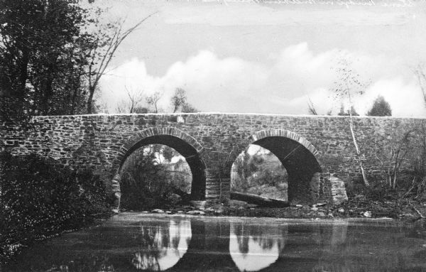 View across water toward a stone bridge with two arches, built in the late nineteenth century, in the wooded setting of Bull Run Battlefield.