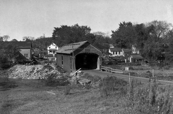 Slightly elevated view across field toward a covered bridge, with town residential buildings in the background.