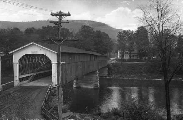 Elevated view of a covered bridge spanning the Delaware River. Signs on the bridge read, "$25.00 Fine for Riding or Driving Faster than a Walk on this Bridge," and "Post No Bills." Two boys are fishing along the riverbank.