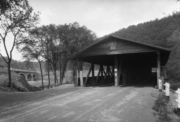View of an older covered bridge in the foreground, with a new bridge in the background on the left. A sign on the covered bridge reads, "McKeesport Automobile Club."