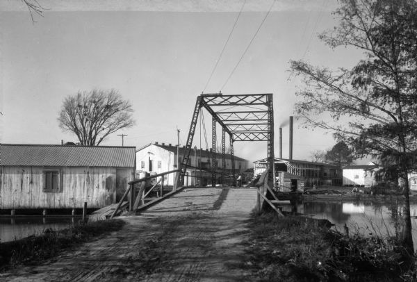 View of Brown's Sawmill and bridge. A man stands in the center of the bridge, and industrial buildings are in the background.