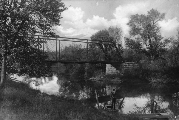 View from shoreline toward Harlem Bridge, a rural bridge over the Pecatonica River. A man is standing in a boat near the riverbank.