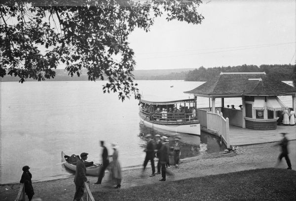 Elevated view of people walking along the shore of Lake Chaubunagungamaug (also known as Webster Lake) in Beacon Park while an excursion boat waits at the dock.