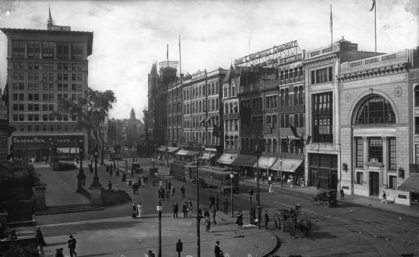 Elevated view of a city square, taken at the end of the nineteenth century. People, carriages, automobiles, and streetcars are in the street. The Merchants National Bank is on the right. Near the building stands a store built in 1870 labeled "Smart Clothes," and the Worcester Business Institute, built in 1870.