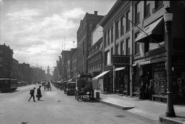 View of a commercial area at the end of the nineteenth century. Men cross the street near parked automobiles, and others stand in the doorway of the Jones Supply Company.