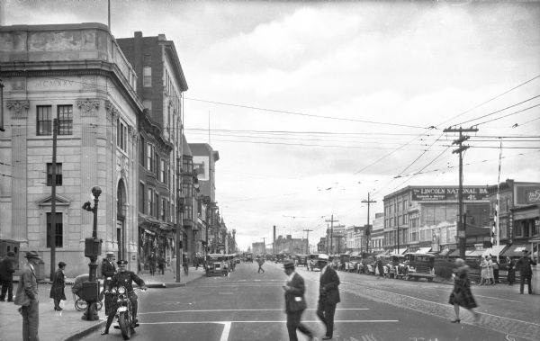 View down Main Avenue, a downtown business district. On the left corner of the avenue stands the Service Trust Company, built in 1925. Men and women cross the street, and a police officer rides a motorcycle on the left.
