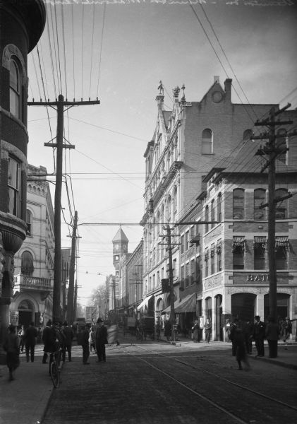View of a crowded intersection at Main Street.  Pedestrians stand on the sidewalk and cross the street near carriages.  On the left corner stands August National Bank, built around 1885.