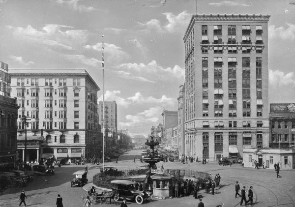 Elevated view of a public square in a business district. Automobiles, pedestrians, and horses and carriages surround a large figural fountain which was erected in 1886.