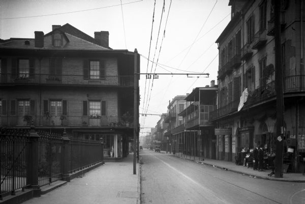 View down sidewalk on left side of Royal Street featuring two men near a market on the right. An iron fence is on the left side of the street.