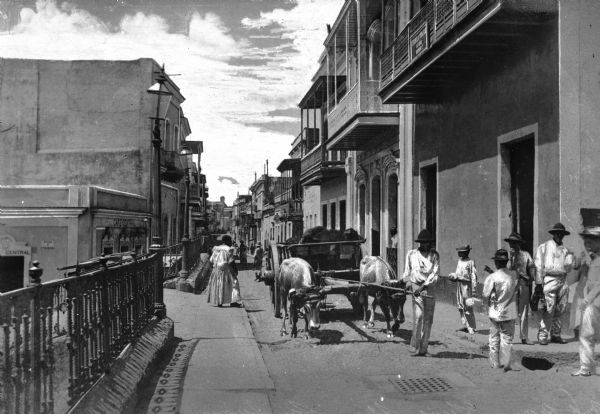 View down Luva Street in San Juan Puerto Rico. Oxen carry a large cart down a narrow street, while people move along the sidewalks.