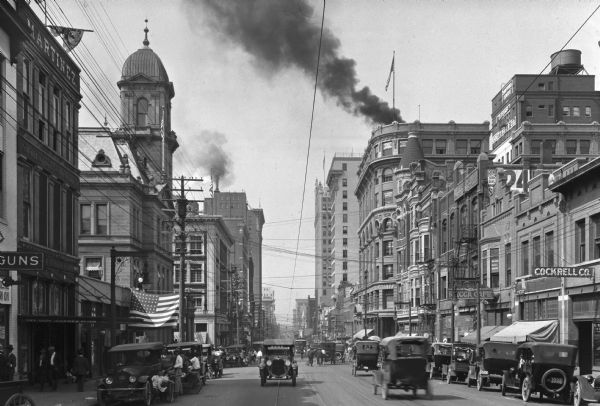 View down street crowded with automobiles and pedestrians. Smoke is billowing from the top of a building on the right.