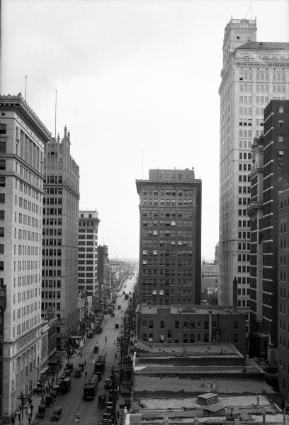 Elevated view of a commercial street with skyscrapers. Below are pedestrians, automobiles, and cable cars.