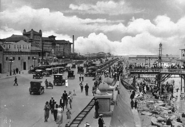 Elevated view down Beach Boulevard, a boardwalk-like street with gift shops and carnival events facing the beach and harbor. Crowds of people are on the sidewalks and the piers.
