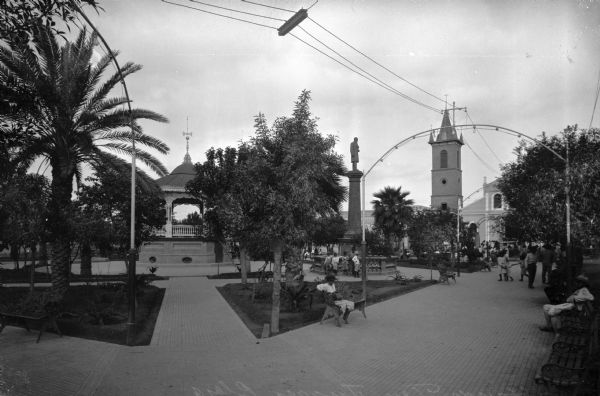 View of Juarez Plaza, a fashionable park square featuring a monument and an elaborately decorated bandstand. People gather on park benches and stand near the monument.