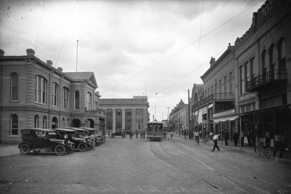 View of Flores Avenue, a commercial section of downtown.  The city hall, built in 1884, stands on the left near Laredo National Bank, founded in 1892.  On the right side of the street, pedestrians gather near the Metropolitan Cafe.