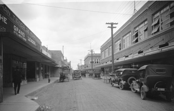 View down a narrow town street lined with buildings, and on the street automobiles and a horse-drawn wagon. A man walks near a clothing store on the left.