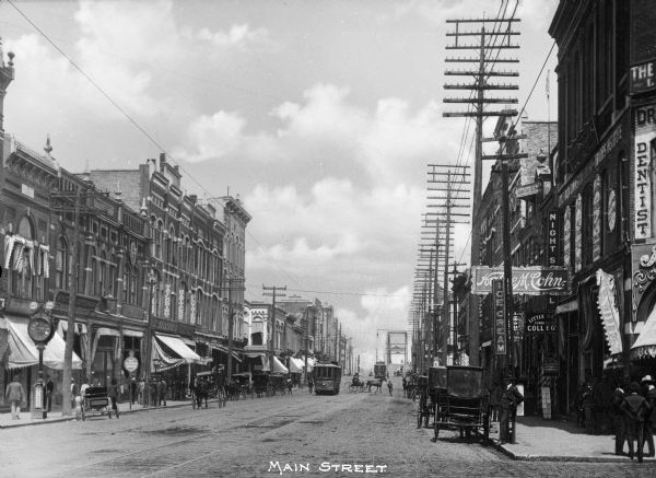 View down Main Street featuring horse-drawn carriages, street cars, and storefronts. The first true non-railway bridge stands in the distance; the 'Free Bridge' spanned the Arkansas River from Main Street to Maple Street, and was completed in 1897. Caption at bottom reads: "Main Street."