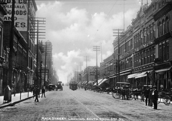 View down Main Street. Pedestrians gather near parked streetcars, horse-drawn carriages, and storefronts. Signs on buildings read: "Pfeifer," "Blass," and "M.M. Cohn & Co." Caption at bottom reads: "Main Street, Looking South From 3rd St."