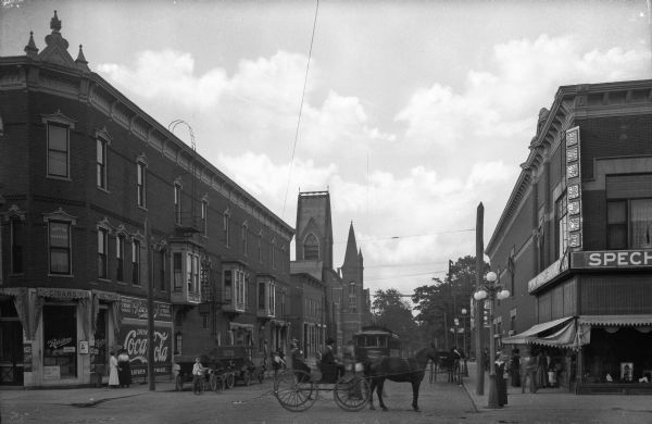 View of a business district featuring horse-drawn carriages and pedestrians near storefronts. On the left corner pedestrians gather near a building labeled "Raisler's Ice Cream," built in 1887.