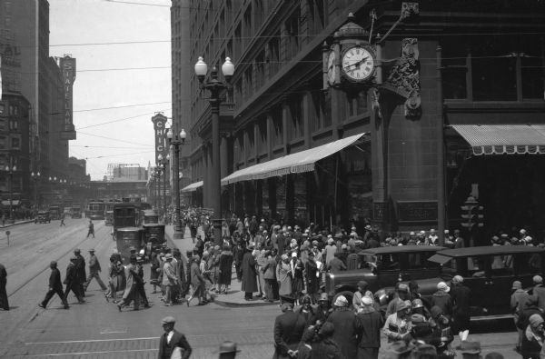 Slightly elevated view of pedestrians and automobiles passing by the flagship Marshall Field's department store (Marshall Field & Co.), built in 1892 by Daniel H. Burnham and located on the corner of State and Washington Streets in the downtown Loop. In the distance is the sign for the Chicago Theater, which opened on October 26, 1921.
