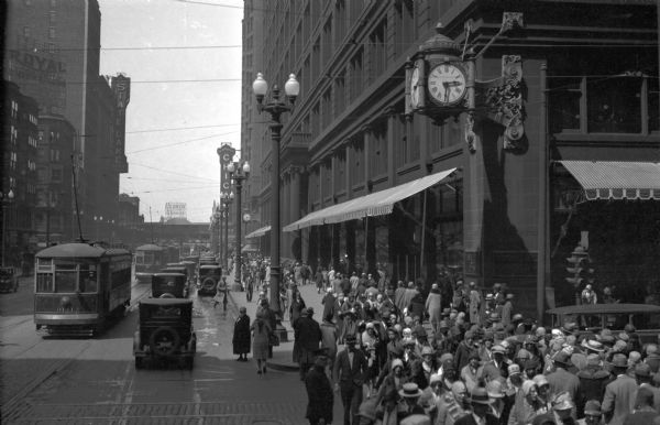 Slightly elevated view of a crowd of pedestrians passing the Marshall Field's department store (Marshall Field & Co.), built in 1892 by Daniel H. Burnham and located on the corner of State and Washington Streets in the downtown Loop. In the distance is the sign for the Chicago Theater, which opened on October 26, 1921.