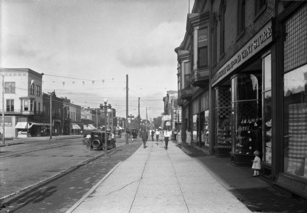 View down sidewalk on right side of a business district featuring automobiles, passengers, and storefronts. On the right a man and child are near the show windows of F.W. Woolworth Co. 5 and 10 Cent Store, founded in 1879.