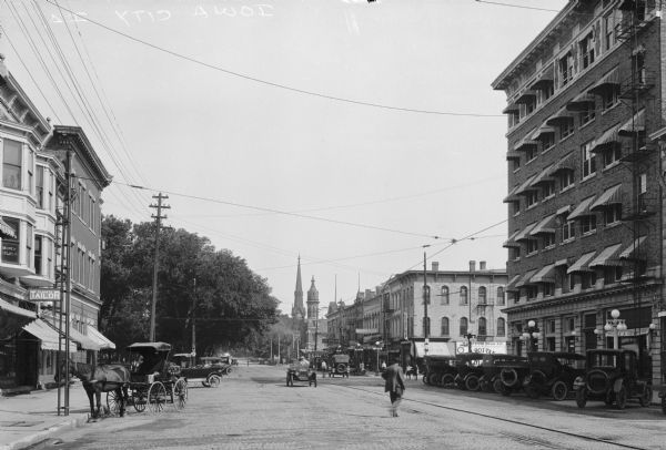 View down a street featuring storefronts, horse-drawn carriages, and automobiles. The steeple of the First Congregational Church, a Gothic Revival building erected in 1869, is in the distance.