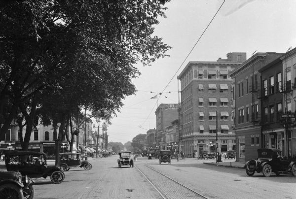 View down a street in a business district. Automobiles are parked at the curb under trees on the left. Along the street are storefronts and the Hotel Jefferson, built in 1913 with a Classical Revival influence.