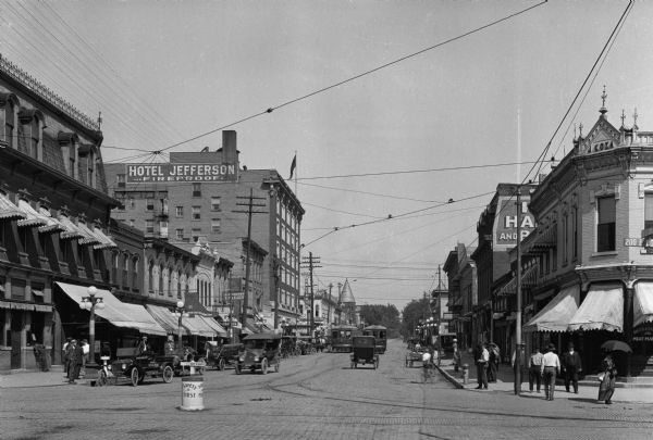 View down South Dubuque Street featuring Hotel Jefferson in the distance at left, built in 1913 with a Classical Revival influence. Automobiles, cable cars, and horse-drawn carriages are along the street outside storefronts.
