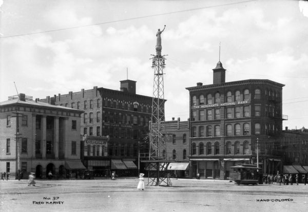 View of the Gottfried Tower, erected in 1895. A woman walks by the iron tower which features a white Statue of Liberty on top and a band platform ten feet above the base. Merchants National Bank is behind the tower; the bank was established in 1879.