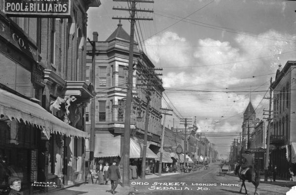 View looking north down Ohio Street. Pedestrians walk by storefronts as a man rides on horseback. Caption reads: "Ohio Street, Looking North, Sedalia, MO."