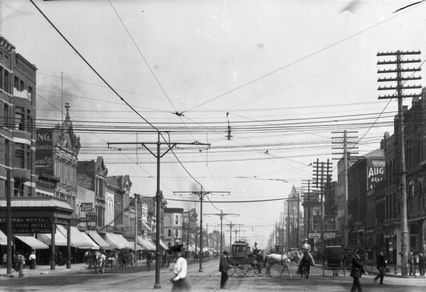 View of a downtown street featuring cable cars, horse-drawn carriages, pedestrians, and storefronts. National Hotel, built in 1855, stands on the street corner at left.
