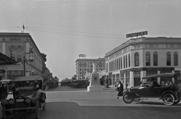 View down a street in a business district. A statue has a sign that reads: "If you can't fight, your dollars can," stands near an intersection. The First National Bank, founded by Robert McHenry in the late 1800s, is on the left corner. Hotel Hughson, built in 1914, is in the distance.