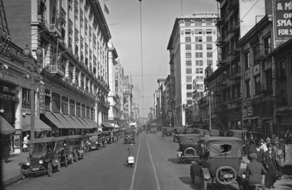 Slightly elevated view down center of Broadway at Eighth Street. Pedestrians cross the street in front of automobiles and cable cars, while stores, theaters, and other tall buildings line the street.