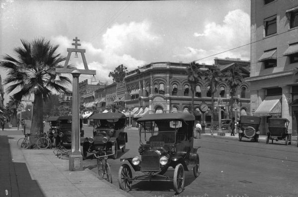 Street Scene featuring, bicycles, automobiles, and palm trees. A large clock stands on a street corner where pedestrians cross, and stone and brick buildings are along the sidewalk.