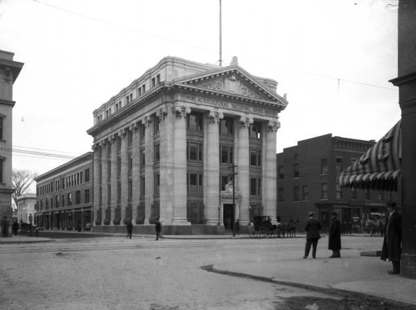 View across intersection toward the Agricultural National Bank, a Neo-Classical structure. Built in 1818 and rededicated in 1908, the bank features an entrance flanked by large columns. Pedestrians and horse-drawn carriages pass by the building.
