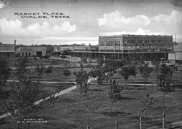 Elevated view of a market place featuring stores and offices. In the foreground is a trail through a group of trees on a lawn that leads to the J.G. Smyth and Company Department Store. Caption reads: "Market Place, Uvalde, Texas."
