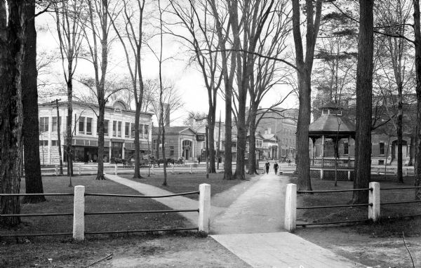 Pedestrians walk through a park past a bandstand and trees. The Park Building can be seen behind them, built in 1914. To its right stands Lee Savings Bank.