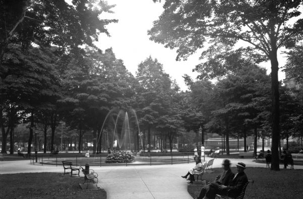 Adults and children sit on park benches surrounding a fountain.