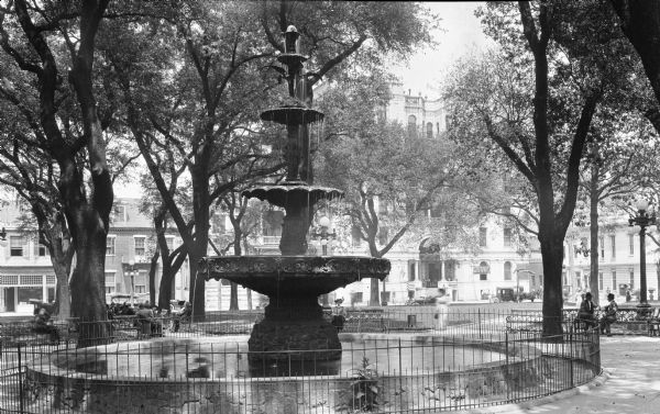 View of Bienville Park. Men and women sit on park benches around a cast iron fountain with an acanthus leaf motif, added to the park in the 1890's. Buildings are in the background behind trees.
