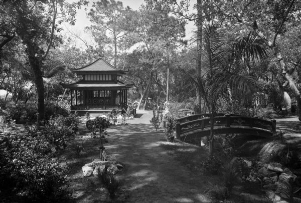 View of the Eagles Nest, a Japanese Garden featuring a bridge and building amongst plants and trees.