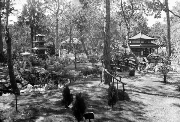 View of the Eagles Nest, a Japanese Garden featuring a bench in the foreground, and beyond a bridge and building amongst plants and trees.