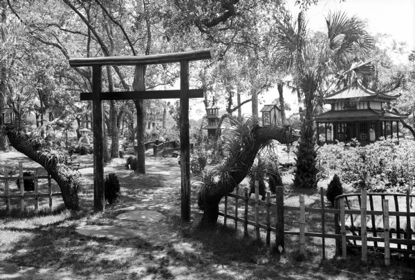 View of the Eagles Nest, a Japanese Garden featuring a wooden Japanese gate, and beyond a bridge and building amongst plants and trees.
