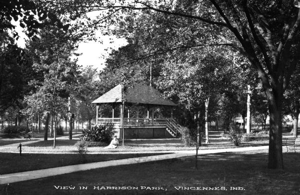 A woman sits in the grass near a bandstand in Harrison Park. Caption reads: "View in Harrison Park, Vincennes, Ind."