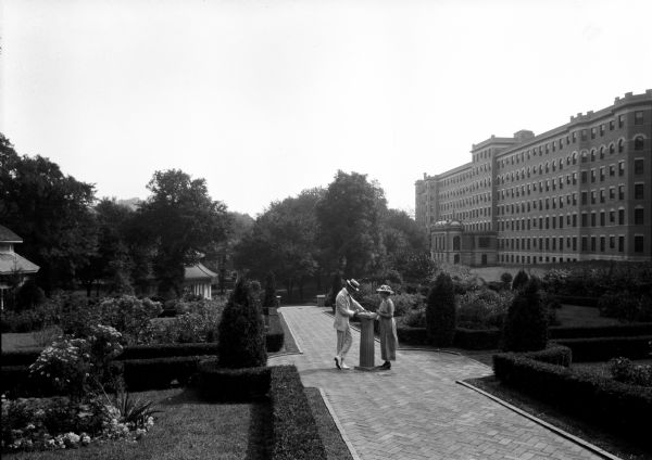 A couple poses near a sundial on a brick walkway in a park. On the right is the original east wing of French Lick Springs Hotel, established in 1845 and built in 1901, and which features an elaborate, colonnaded entrance.