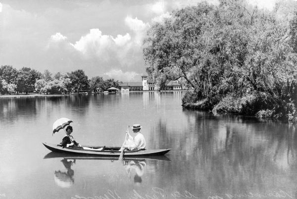 View of a canoe outing in the city park. A woman reclines, holding an umbrella, and a man holds an oar, paddling.