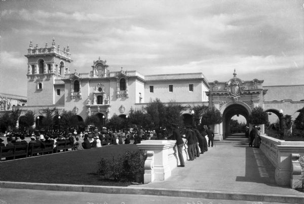 A crowd gathers in Balboa Park to listen to a band performance. The park stands in front of the Foreign Arts Building, constructed in 1915.